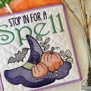 May include: A white quilted fabric square with purple, green, and orange embroidery. The text "Stop in for a Spell" is embroidered in green. The design includes a witch's hat, bats, and pumpkins.