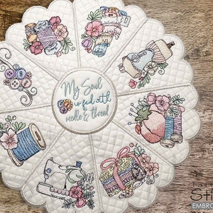 Puede incluir: Un cubremesa redondo acolchado blanco con borde festoneado. El centro presenta el texto "My Soul is fed with needle & thread". El cubremesa está decorado con varios diseños de temática de costura, incluyendo carretes de hilo, botones y una máquina de coser.