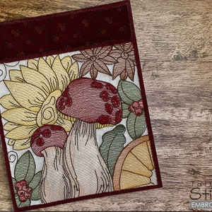 Könnte beinhalten: Bestickte Notizbuchhülle mit einem Design aus Pilzen, Sonnenblumen und Blättern. Die Hülle hat eine bordeauxfarbene Oberseite und einen weißen Hintergrund mit detaillierten Stickereien in verschiedenen Farben. Das Design enthält den Text "Tattered Stitch Embroideries".