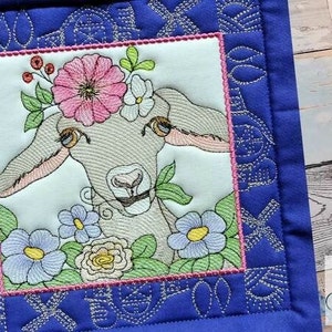 Peut inclure: Broderie représentant une chèvre portant une couronne de fleurs roses, blanches et jaunes. La chèvre est entourée de fleurs bleues, roses et blanches. Le motif est encadré dans un carré bleu avec une bordure argentée.