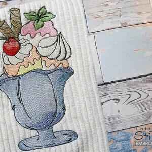 Puede incluir: Toalla blanca bordada con un diseño de helado colorido. El helado incluye helado rosa y blanco, una cereza roja y barquillos, todo en una copa azul. La toalla tiene una textura acanalada.