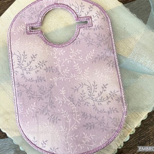 May include: A lavender embroidered bib with a floral pattern and a decorative border. The bib has a circular opening at the top and is placed on a light-coloured surface with a blue ribbon.