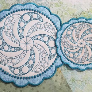 May include: Two teal blue embroidered coasters with a white and teal blue geometric design. The coasters have a scalloped edge.