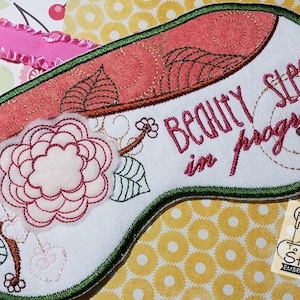 May include: A white sleep mask with a green border and a pink floral design. The text "Beauty sleep in progress" is embroidered on the mask.