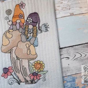 May include: Embroidered design of two gnomes sitting on top of mushrooms with flowers and leaves around them. The gnomes are wearing hats and have long hair. The mushrooms are brown and white.