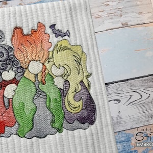 Könnte beinhalten: Gestickte Darstellung von drei Hexen mit lila, grüner und roter Kleidung. Die Hexen haben orange, gelbes und lila Haar. Das Design beinhaltet eine Fledermaus.