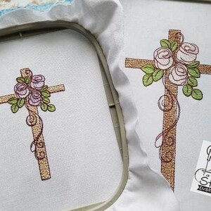May include: Two embroidered designs of a wooden cross with a floral arrangement of pink roses and green leaves. The cross is brown and the roses are pink with a hint of purple. The designs are on white fabric.
