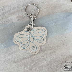 Può includere: Un portachiavi in ​​pelle bianca con un design a farfalla ricamato in blu. Il portachiavi ha un anello in metallo argentato e una chiusura in metallo argentato.