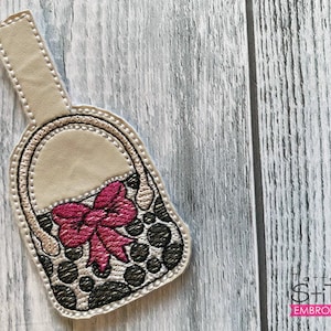 Puede incluir: Un llavero de cuero beige con un diseño de lunares negros y blancos y un lazo rosa. El llavero tiene la forma de un pequeño bolso de mano.