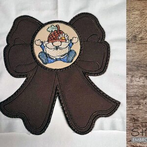 May include: Embroidered brown ribbon with a cartoon gnome in a circle. The gnome wears a hat and blue outfit. The ribbon has a black stitched outline and is on a white background.