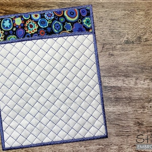 May include: A white quilted fabric square with a blue and purple patterned border. The fabric is quilted in a diamond pattern.