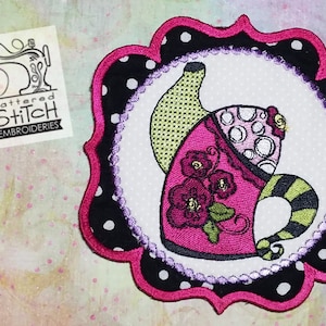 May include: A pink and green teapot with a floral design, surrounded by a black and white polka dot border. The teapot is embroidered on a white fabric with a scalloped edge.