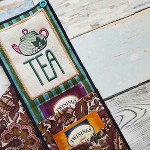 May include: A teal, green, and brown fabric tea bag organizer with a pocket for tea bags and a pocket with an embroidered teapot and the word "TEA".