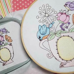 May include: Embroidered floral design featuring a teacup overflowing with flowers and birds in a variety of colors. The embroidery is set in a wooden hoop and a square frame. The design includes pink, orange, blue, and purple elements.