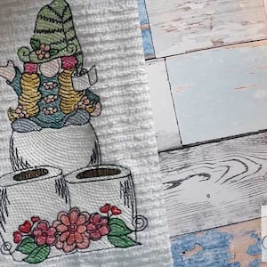 Puede incluir: Diseño bordado de un gnomo sosteniendo un rollo de papel higiénico, de pie sobre dos rollos de papel higiénico con flores rosas y hojas verdes.