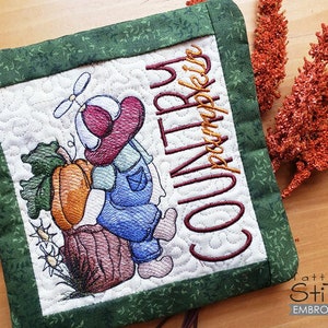 May include: A green and white quilted pot holder with a country pumpkin design. The design features a cartoon character holding a pumpkin and the text "Country Pumpkin".