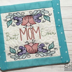 May include: Square quilted pot holder with a turquoise border and white polka dots. The center features embroidered flowers and the text "Best MOM Ever" in a decorative font. A loop is attached for hanging.