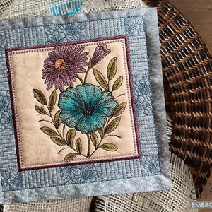 May include: A square quilted fabric panel with a floral embroidery design. The embroidery features three flowers in shades of purple, blue, and green. The panel is framed with a blue and white quilted border.