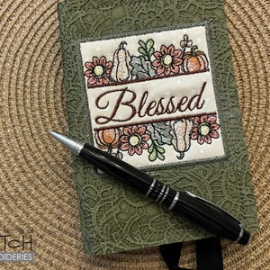 Puede incluir: Un diario verde con un diseño bordado de flores y calabazas, la palabra "Blessed" está bordada en el centro. Un bolígrafo negro con detalles plateados descansa sobre el diario, que está sobre una superficie tejida.