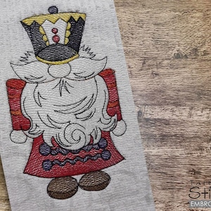 May include: Embroidered design of a red and white nutcracker with a black hat and a white beard. The nutcracker is holding a red and white staff.