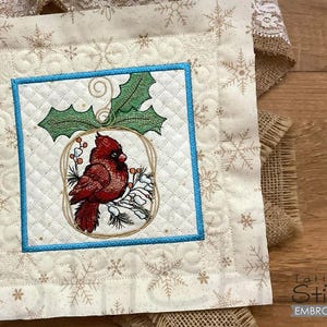 May include: A white quilted fabric square with a blue border, featuring an embroidered design of a cardinal perched on a holly branch with red berries. The background is a cream color with a snowflake pattern.