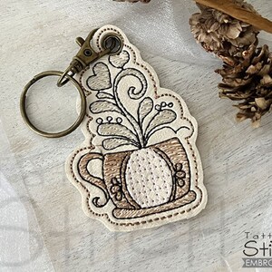 Könnte beinhalten: Bestickter Schlüsselanhänger mit einem Kaffeebecher-Design und einer Pflanze. Der Schlüsselanhänger hat einen bronzefarbenen Karabiner und Schlüsselring. Das Design ist in Beige- und Brauntönen gehalten und hat eine gepunktete Kontur. Der Schlüsselanhänger ist von Tattered Stitch Embroideries.