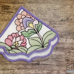 Puede incluir: Un diseño de esquina bordado en morado y blanco con un motivo floral. El diseño presenta flores rosas, amarillas y verdes con un borde festoneado.