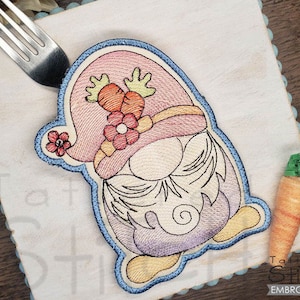 May include: An embroidered Easter gnome with a pink hat, carrot details, and a flower accent. The gnome is outlined in blue and sits on a white square with a fork. A carrot ornament hangs nearby. The image is from Tattered Stitch Embroideries.