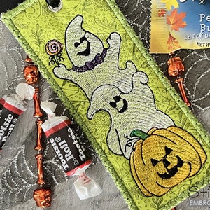 Może przedstawiać: Zielona zakładka z tkaniny z wyszytymi obrazami dyni i dwóch duchów. Zakładka ma pętlę u góry do przymocowania do książki. Tekst "Tootsie Roll®" jest widoczny na opakowaniu cukierków na zdjęciu.