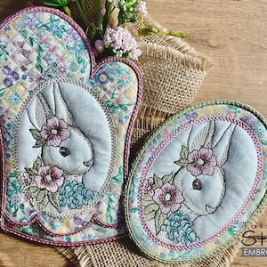 Może przedstawiać: Dwa pikowane uchwyty do garnków z haftowanymi twarzami królików i wzorami kwiatowymi. Uchwyty do garnków są w odcieniach różowego, niebieskiego, zielonego i żółtego. Większy uchwyt do garnka ma kształt rękawicy, a mniejszy jest owalny.