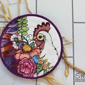以下が含まれることがあります： カラフルな花の模様が施された雄鶏の刺繍デザイン。雄鶏は白く、赤い鶏冠と肉垂れがあります。花はピンク、紫、黄色です。デザインは丸い紫色の縁取りのある布地です。