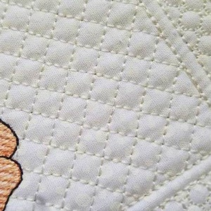 May include: Close-up of a quilted fabric with a repeating diamond pattern in off-white. A portion of an embroidered flower in shades of peach and yellow is visible. The logo for Tattered Stitch Embroideries is in the bottom right corner.