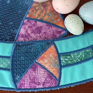 Puede incluir: Un calentador de taza de café de tela azul turquesa con un diseño de patchwork de tela azul, morado, naranja y verde. El calentador tiene un botón azul en el lateral.