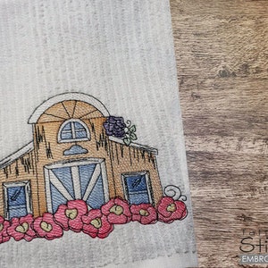 Puede incluir: Diseño bordado de un granero marrón con una puerta y ventanas azules, rodeado de flores rosas. El granero tiene un techo blanco y una pequeña ventana en la parte delantera.