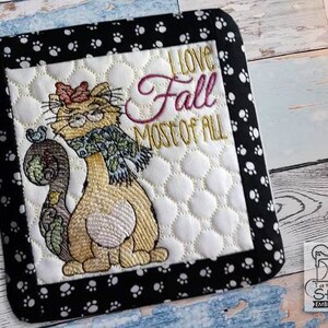 May include: Embroidered kitchen pot holder with a black and white paw print border. The center features a cartoon cat wearing a scarf and the text "I love Fall most of all".