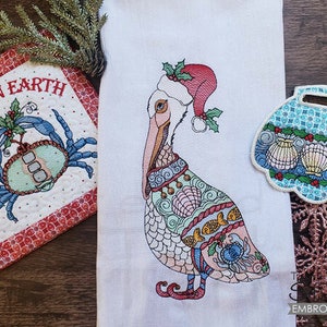 May include: A white kitchen towel with a colorful embroidered pelican wearing a Santa hat. The pelican is decorated with seashells, fish, and other nautical designs. The towel also features a small embroidered seashell ornament with holly and berries.