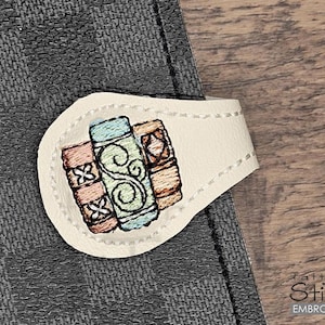 May include: A white leather bookmark with an embroidered design of three stacked books. The books are decorated with colorful swirls and patterns.