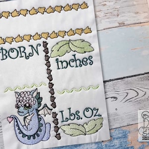May include: Embroidered design featuring a cute blue dragon with a heart, surrounded by green leaves. The text "BORN" and "inches" are above the dragon, and "lbs. oz." is below. The design is perfect for a baby's nursery or a baby shower gift.