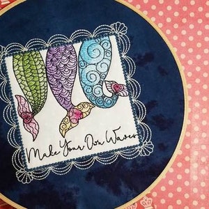 May include: Embroidered design of three mermaid tails in shades of green, purple, and blue with a white background. The design is framed with a white scalloped border and the text "Make Your Own Waves" is stitched below the tails.