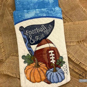 Op de afbeelding: Een gewatteerd stoffen item met een blauw en wit bovenstuk, met een geborduurde voetbal, pompoenen en een banner met de tekst "Football & Fall Y'all". Het ontwerp bevat een bruine voetbal en oranje en blauwe pompoenen.