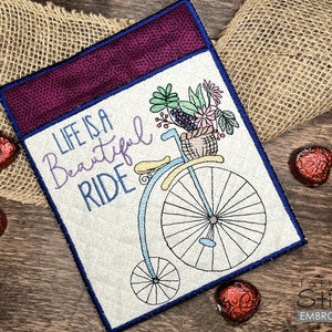 May include: A white embroidered design with a bicycle with a basket of flowers and the text "Life is a Beautiful Ride". The design is on a purple fabric with a white border.