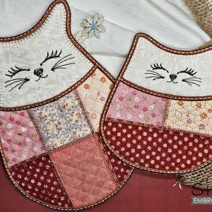 Puede incluir: Dos agarraderas de olla con forma de gato acolchadas con un diseño de patchwork en tonos de rosa, rojo y crema. Los gatos tienen caras bordadas con ojos negros y bigotes.