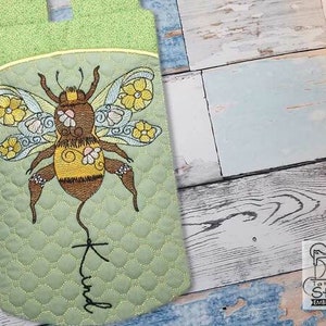 Puede incluir: Una bolsa de tela acolchada verde con una abeja bordada en ella. La abeja tiene alas marrones y amarillas con flores y la palabra "Kind" escrita debajo.