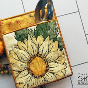 May include: A beige fabric utensil holder with a sunflower design and a gold top. The holder has a pocket for a spoon or other utensil.