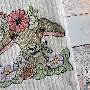 Peut inclure: Serviette blanche brodée avec une tête d'agneau détaillée ornée d'une couronne florale et entourée de fleurs roses, violettes et vertes. Le visage de l'agneau est beige avec des accents roses. La serviette a une texture côtelée.