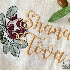 Puede incluir: Diseño bordado con la frase "Shana Tova" en letras doradas con una granada y hojas verdes. El diseño está sobre un fondo de tela blanca.