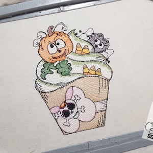 May include: A Halloween-themed cupcake with green frosting, candy corn, a pumpkin face, and a spider on top. The cupcake is decorated with a pink ribbon and a skull.