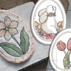 Puede incluir: Tres posavasos ovalados bordados con diseños de temática primaveral. Uno presenta un conejito blanco con flores rosas, otro un ramo de tulipanes rosas y el último una flor amarilla con hojas verdes.