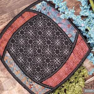 Puede incluir: Un posavasos cuadrado acolchado con un centro negro y un borde multicolor. El borde presenta tonos de azul, rojo y marrón. El posavasos está hecho de tela y tiene un diseño acolchado.