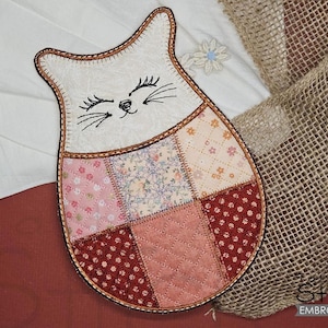 May include: A white cat-shaped quilted hot pad with a patchwork design in shades of pink, red, and cream. The cat has a smiling face with black embroidered whiskers and eyelashes.